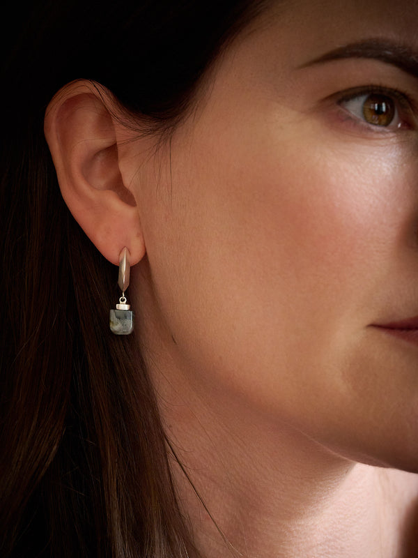 A close-up side profile of a woman wearing a small silver-toned drop earring with a rectangular gray stone pendant against a dark background.