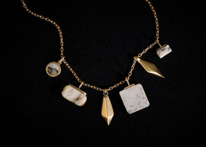 A gold chain necklace featuring multiple organic-shaped and geometric stone pendants in white, gray, and marble stone, displayed against a solid black background.